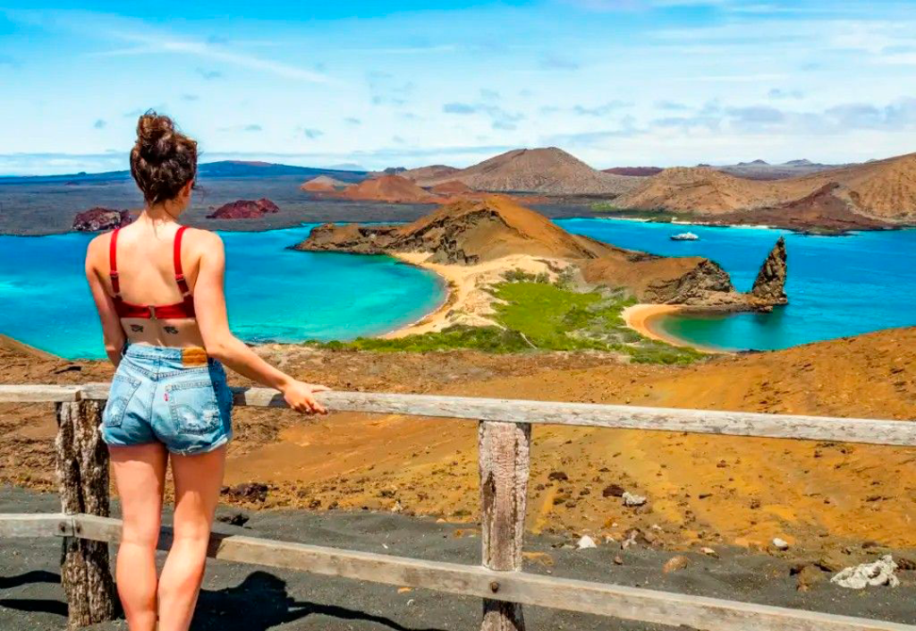 Fotografía de una mujer observando de una parte de las Islas Galápagos donde se aprecia la Punta Pitt junto a la playa Oliviana y al fondo la Isla San Cristóbal